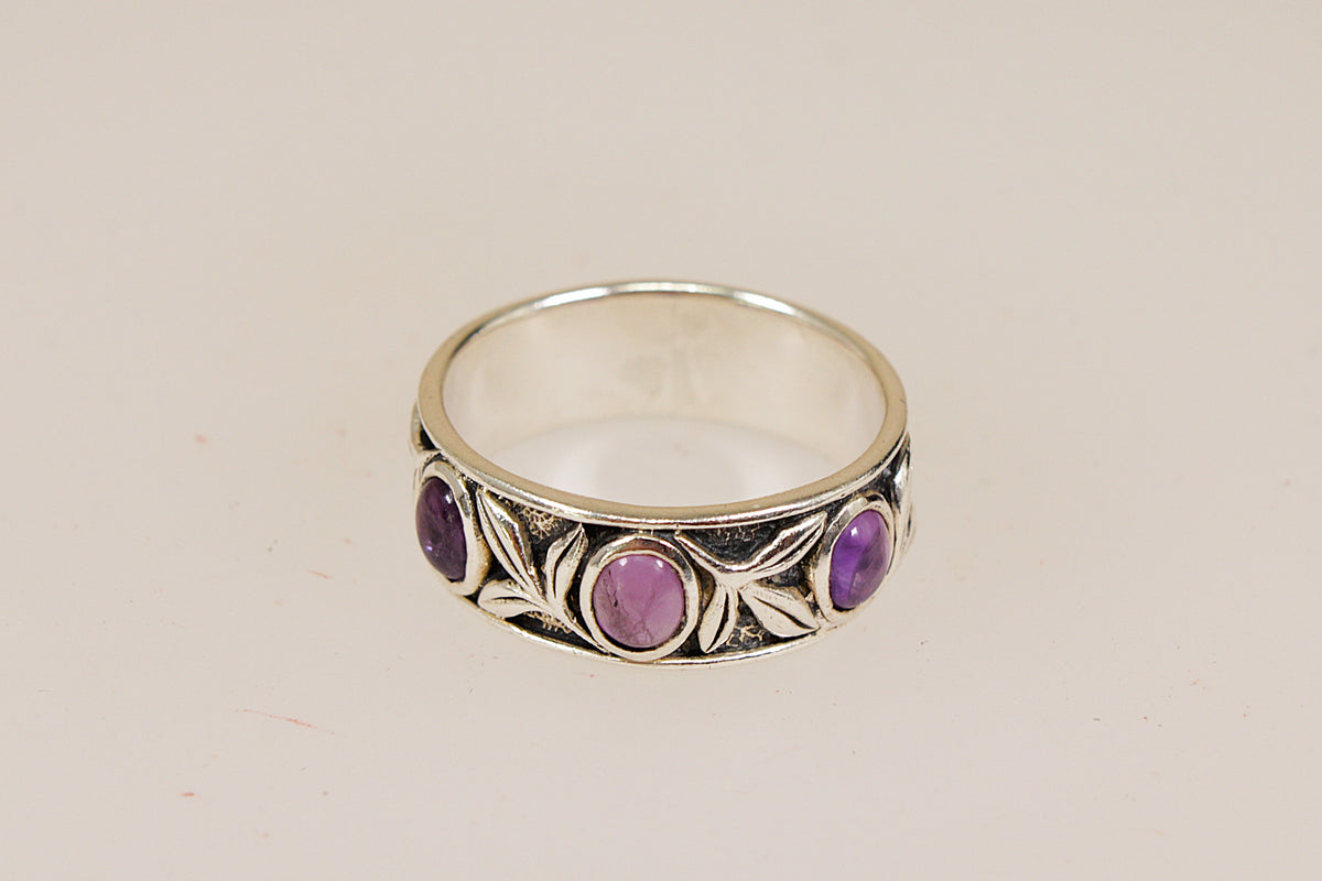 A sterling silver ring featuring a decorative leaf pattern and four oval purple amethyst gemstones, shown on a plain beige background.