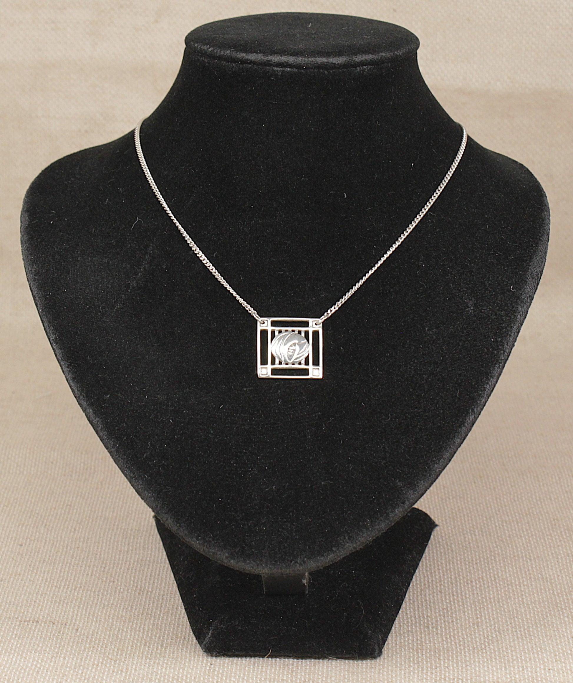 A silver geometric square pendant necklace displayed on a black velvet jewelry bust against a neutral beige background.