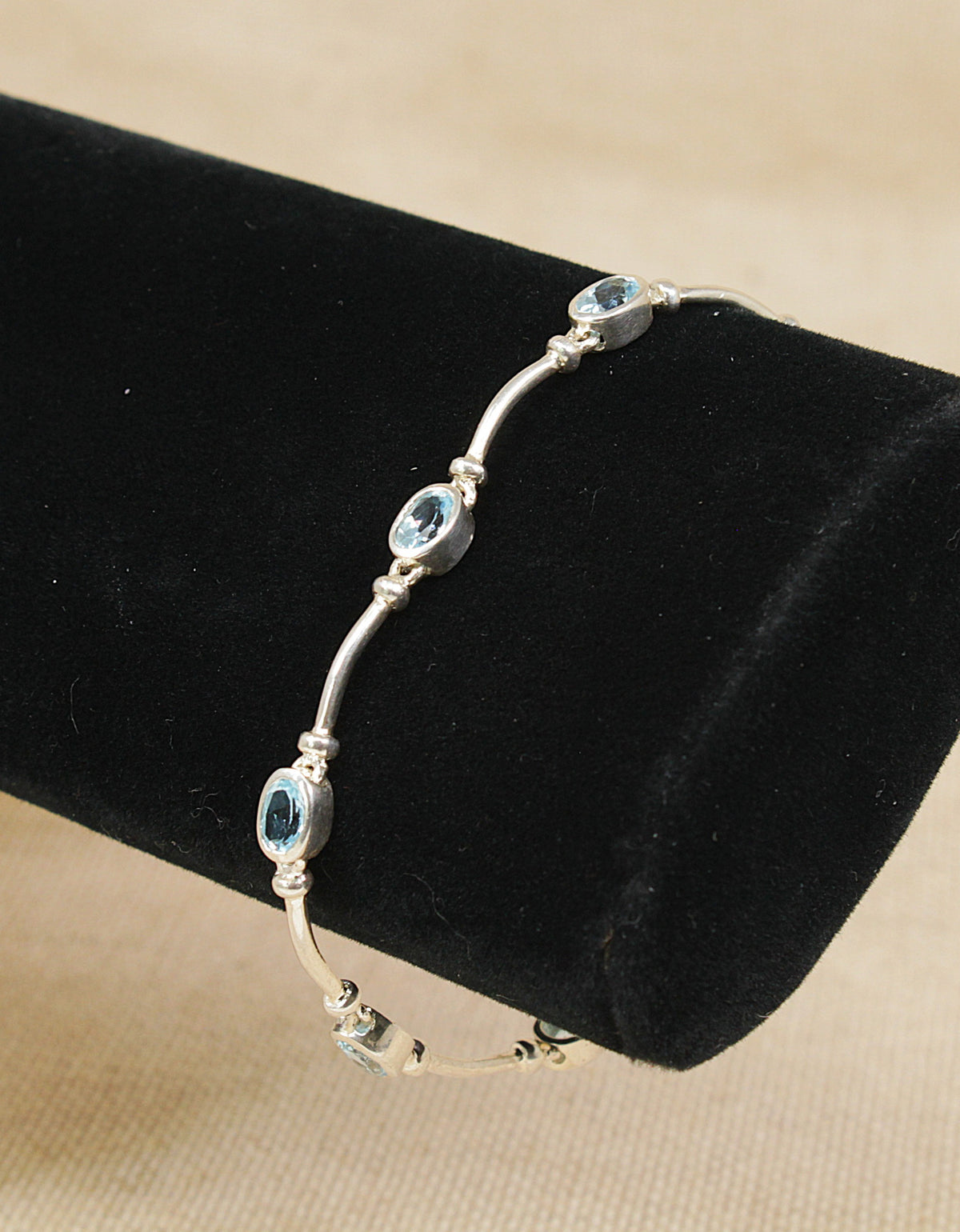 A silver bangle bracelet with three oval light blue topaz gemstones, displayed on a black velvet jewelry stand against a beige fabric background.