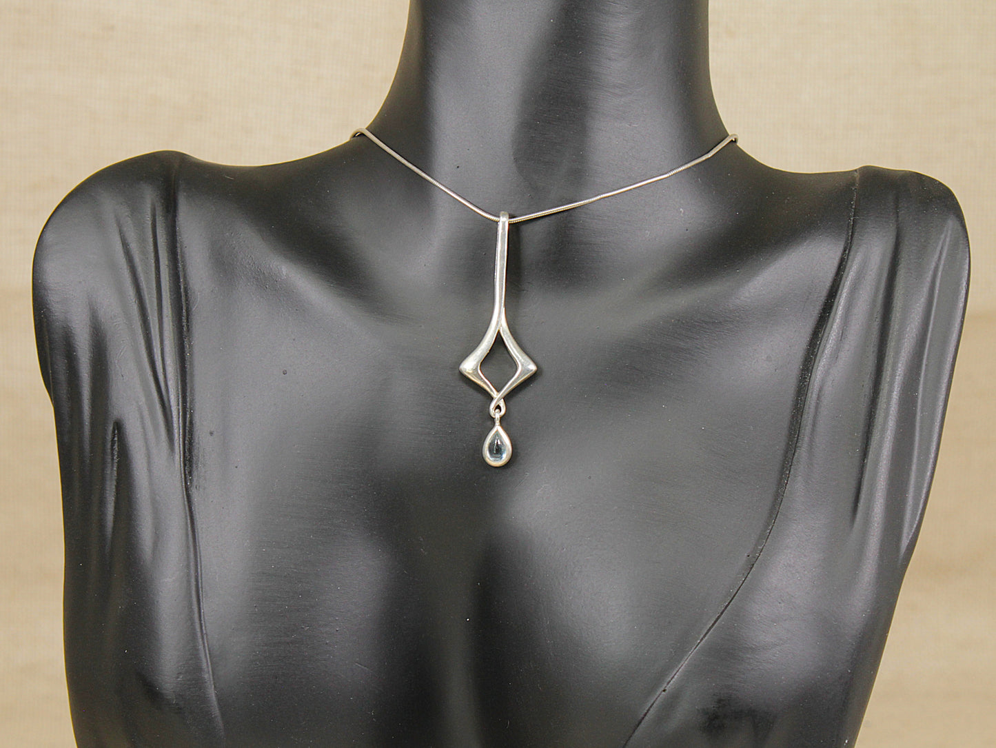 A sterling silver pendant necklace with a teardrop-shaped blue topaz drop and openwork diamond design, displayed on a black mannequin bust against a beige background.