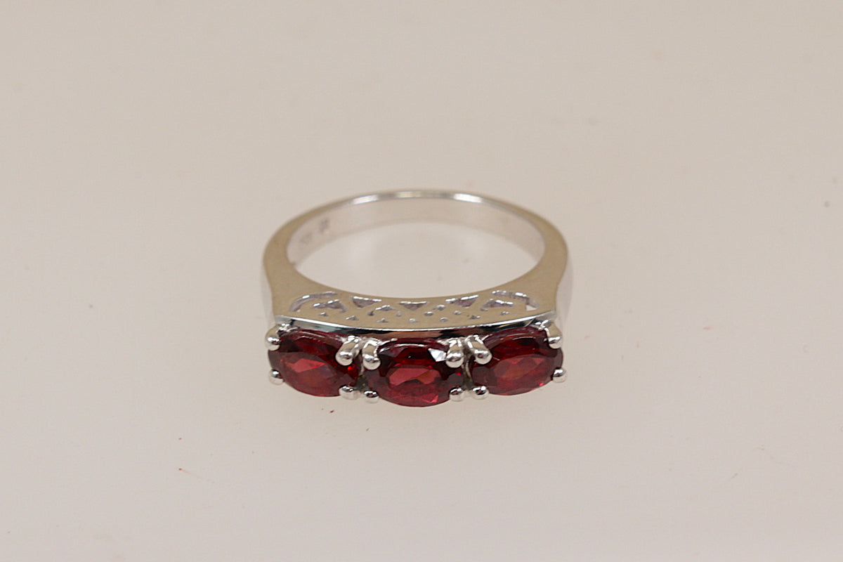 A silver ring featuring three oval-cut deep red garnet gemstones, set against a plain light beige background.