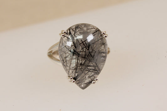 A silver ring featuring a large teardrop-shaped clear quartz gemstone filled with tourmaline barbs, set in a four-prong setting on a plain light beige background.