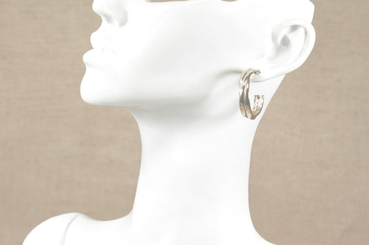 A pair of sterling silver hoop earrings shown on a white mannequin head and neck against a neutral beige background.