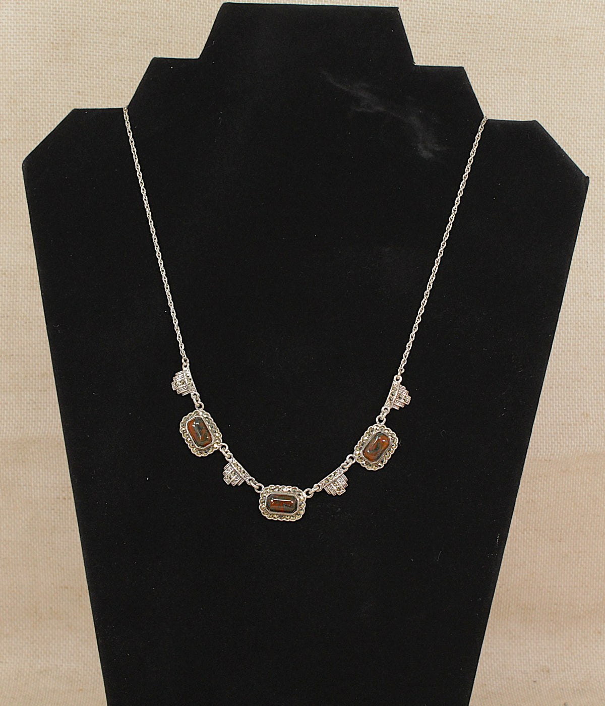 A silver necklace with three rectangular amber  gemstones and small accent marcasite gemstones, displayed on a black jewelry bust against a beige textured background.