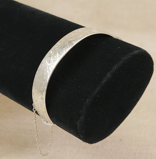 A polished silver bangle bracelet with engraved floral patterns, displayed on a black velvet cylindrical jewelry stand against a beige fabric background.