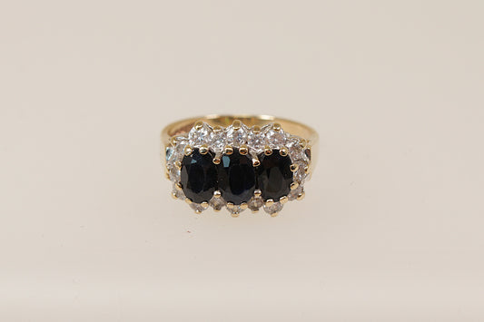 A vintage 9ct gold sapphire trilogy ring with claw set oval blue sapphires and cubic zirconia gemstones around the mount, on a white background.