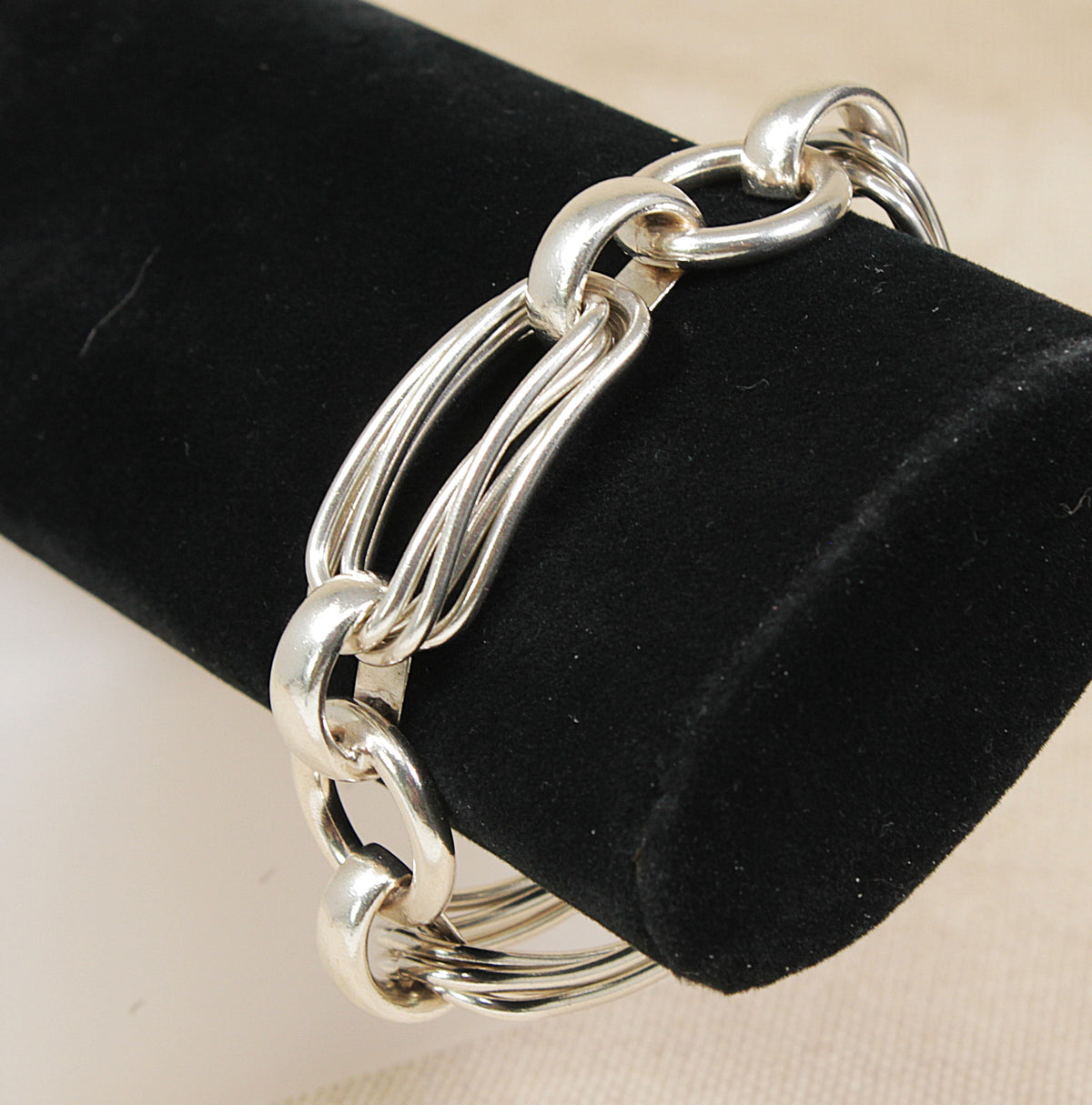 A polished silver-toned chain link bracelet displayed on a black velvet jewelry stand against a light beige background.