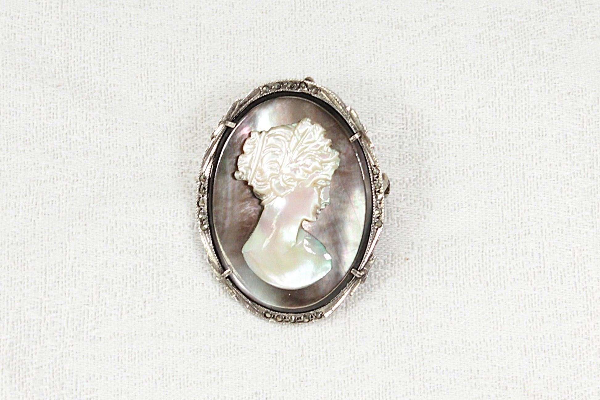 Cameo brooch with a carved mother of pearl woman's face in a silver and marcasite oval frame on a white background
