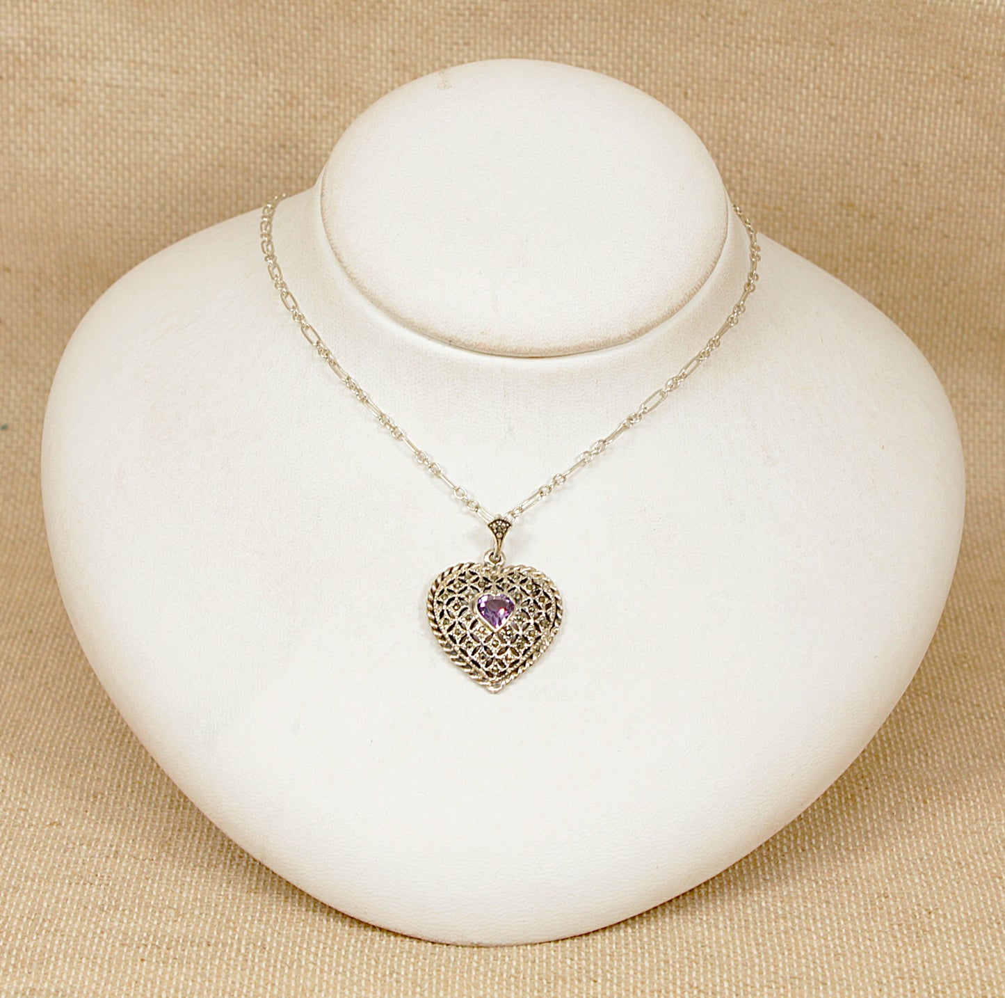 Necklace with a heart-shaped pendant  with a central faceted purple amethyst  on a white stand against a beige background