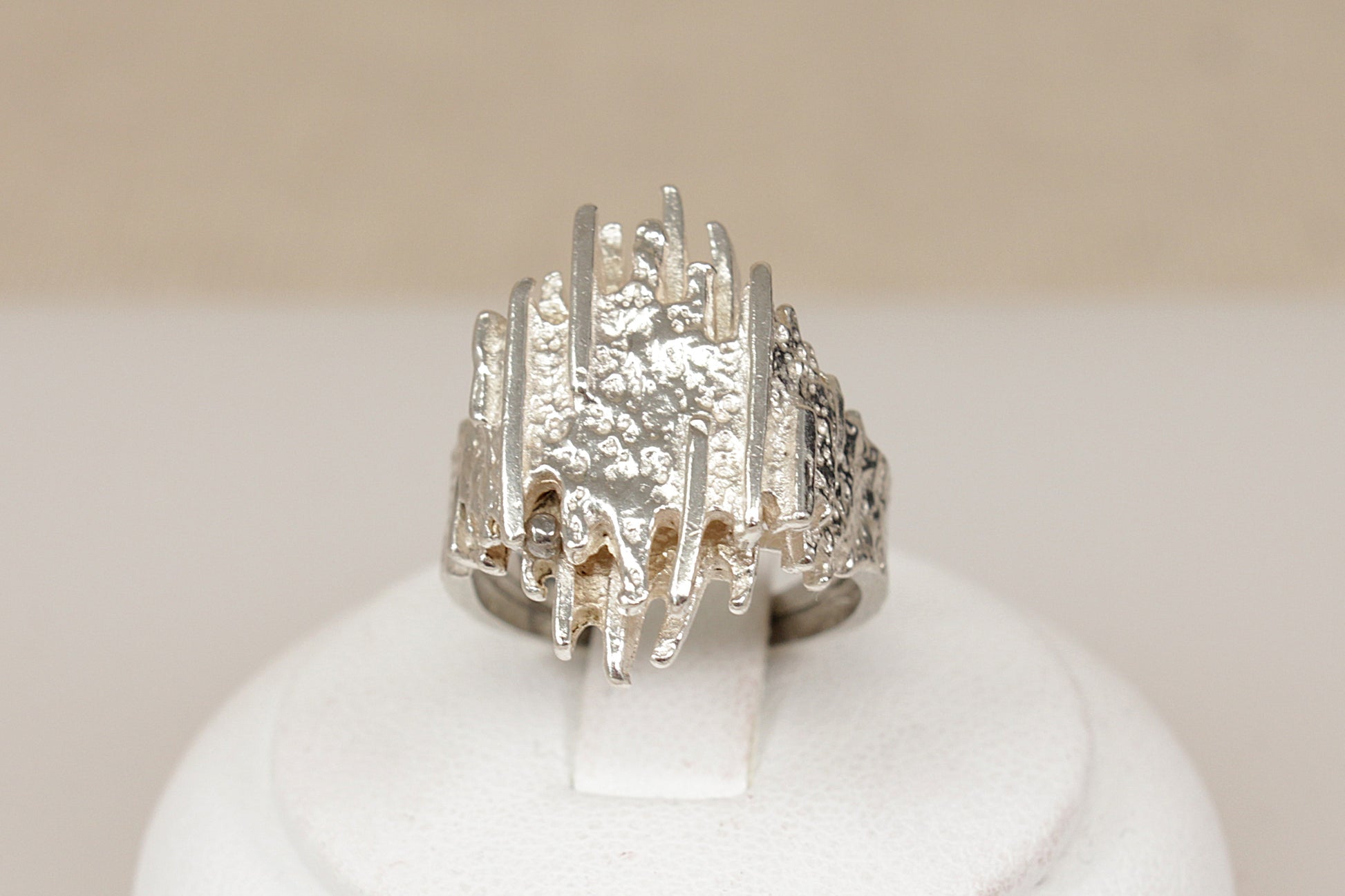 A textured  brutalist sterling silver ring displayed on a white ring stand against a soft beige background.