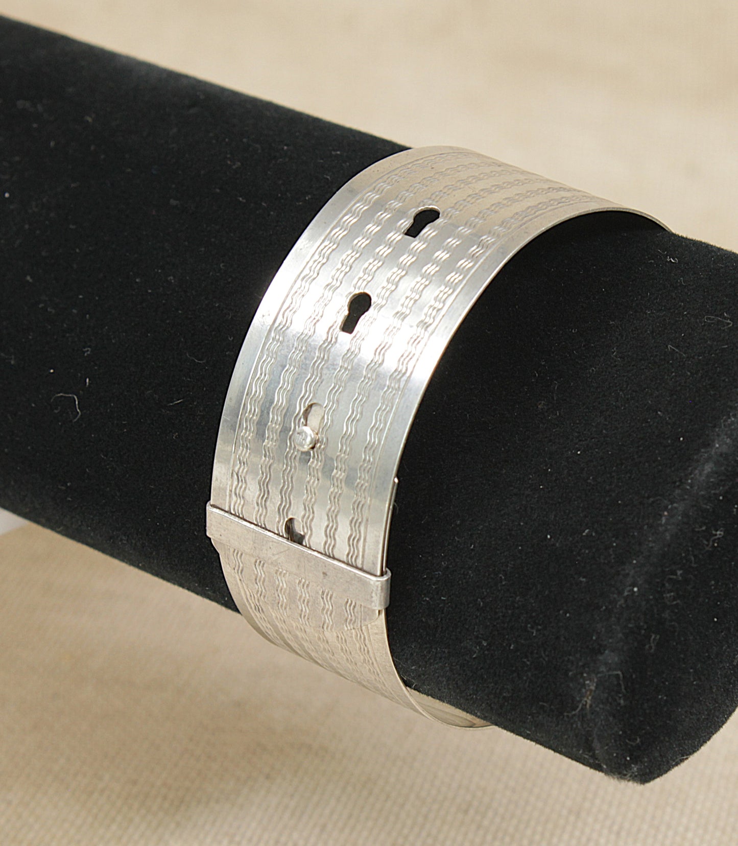 A wide silver cuff bangle bracelet with a textured, hammered pattern and three small cutouts, displayed on a black cylindrical jewelry stand against a light beige fabric background.