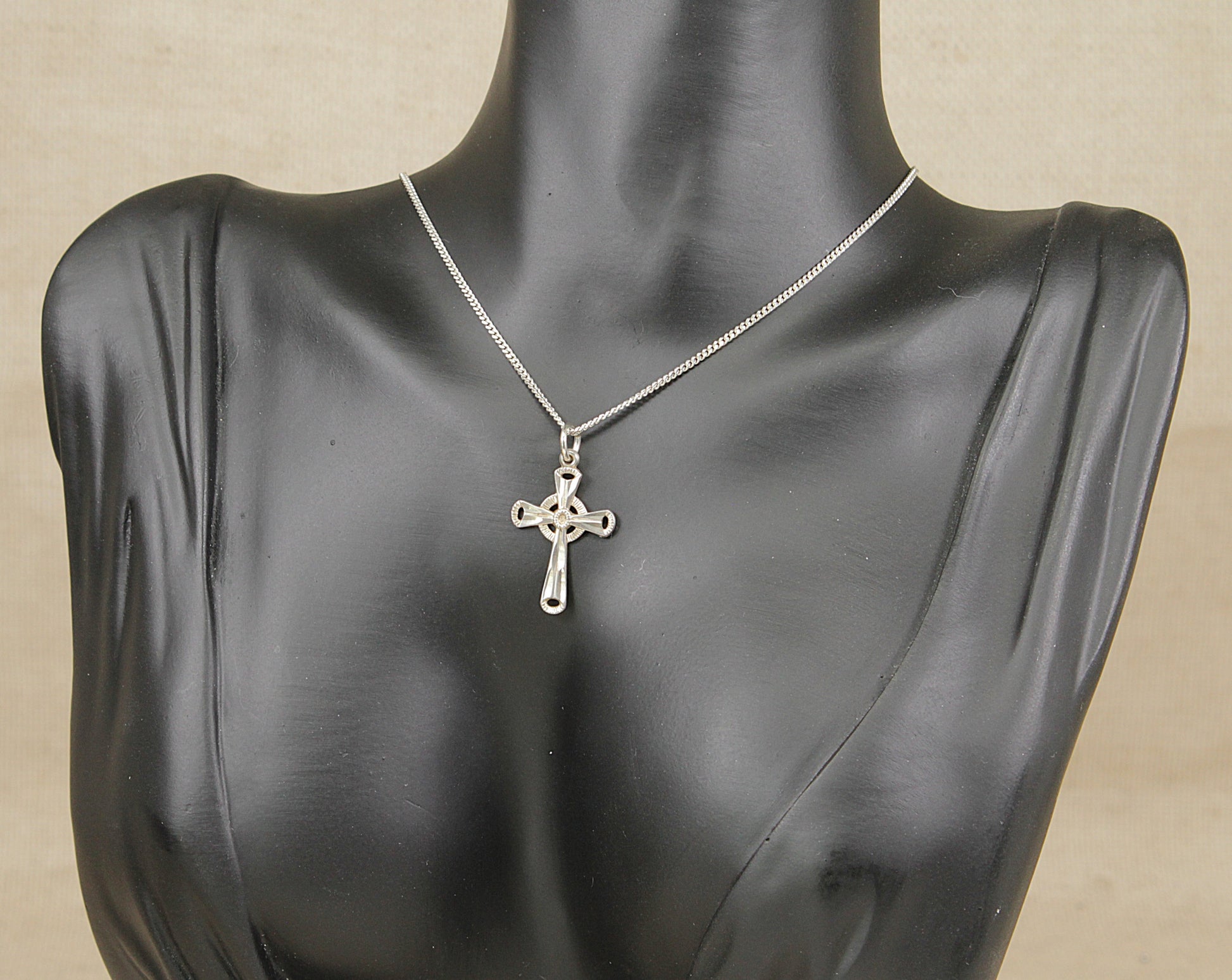 A silver cross pendant necklace on a silver chain, displayed on a black mannequin bust against a light beige background.