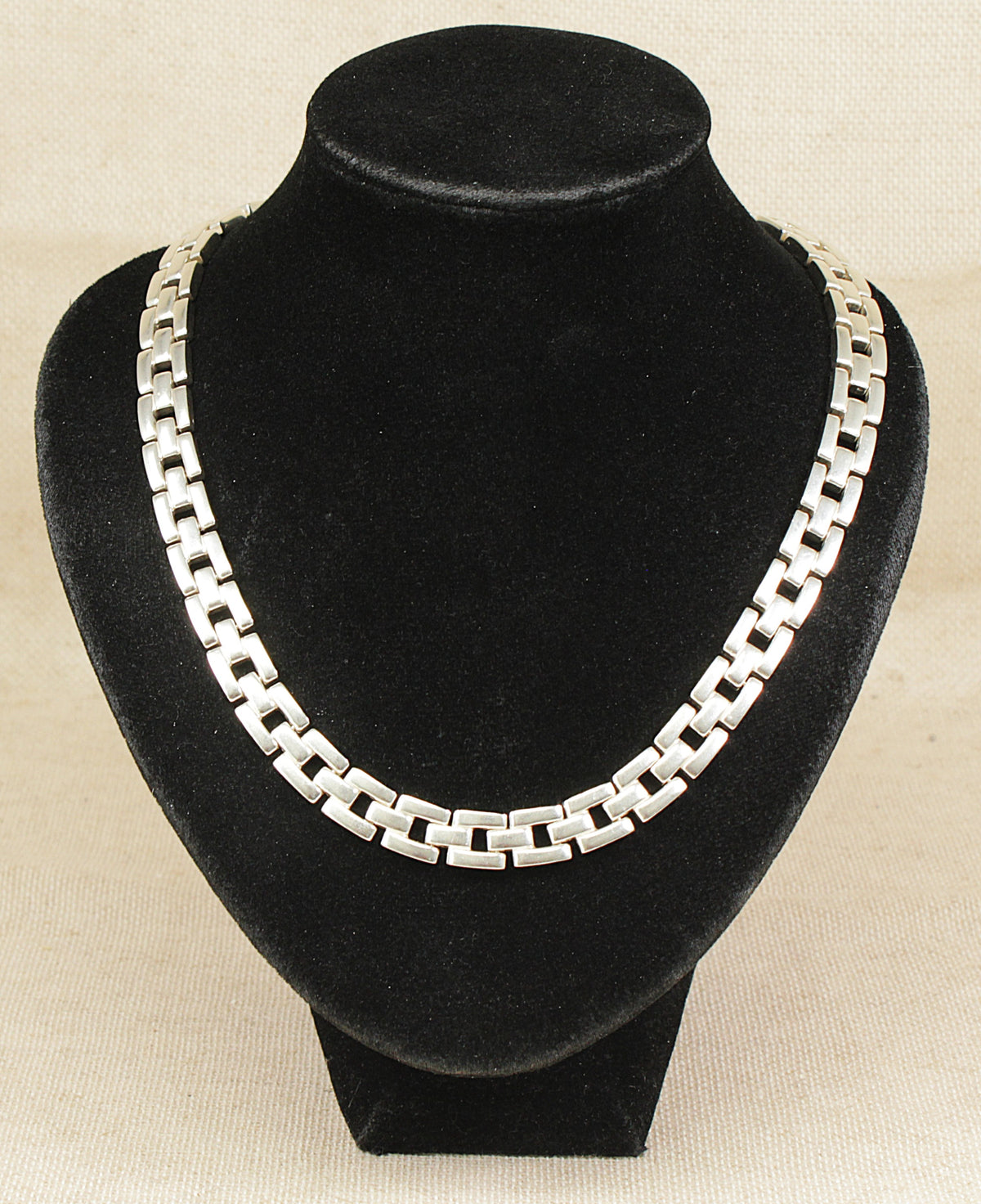 A silver-toned interlocking link chain necklace displayed on a black velvet jewelry bust against a light beige textured background.