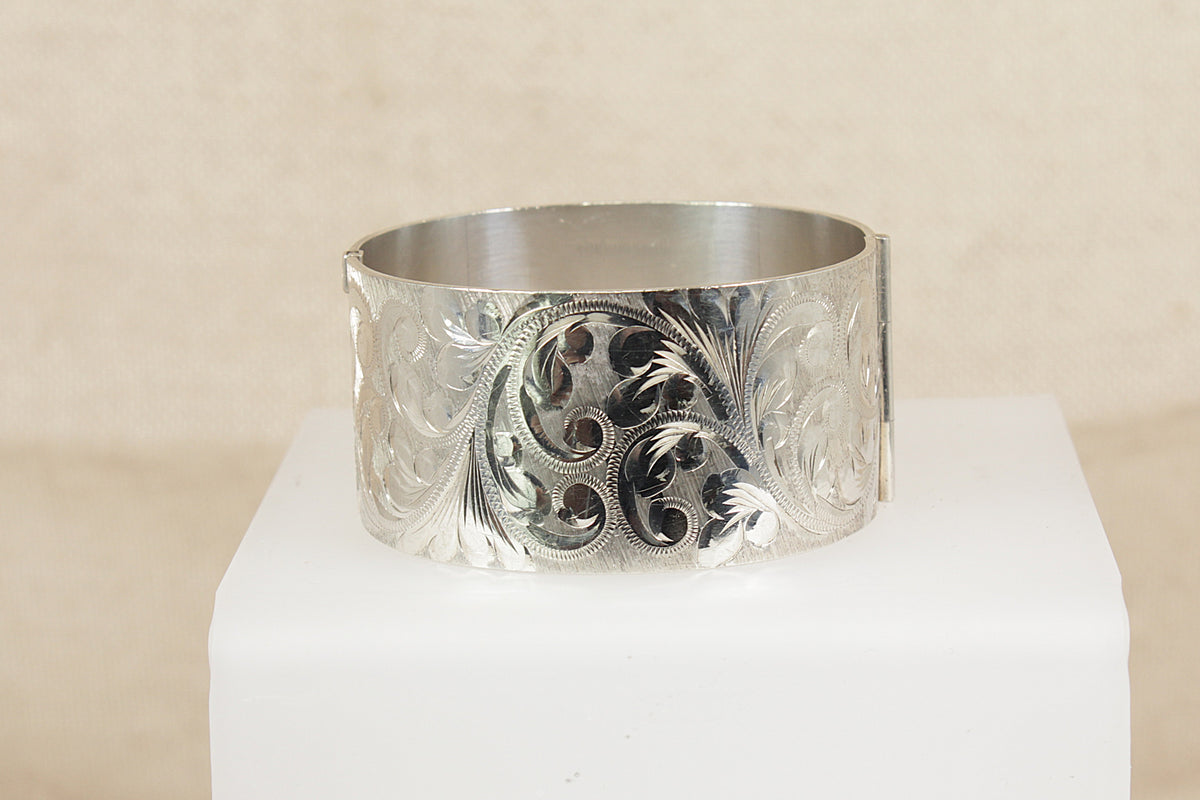 A wide silver cuff bracelet with intricate floral and scroll engraving, displayed on a white block against a neutral beige background.