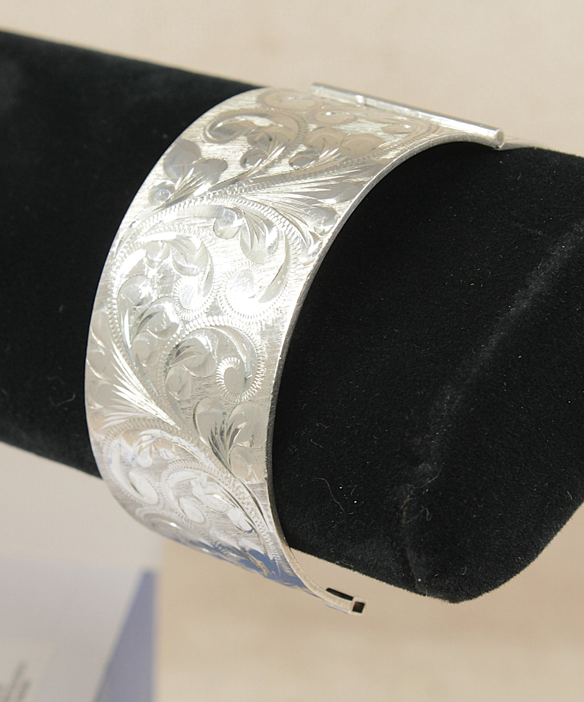 A wide silver cuff bracelet with intricate floral engraving, displayed on a black velvet jewelry stand against a light beige background.