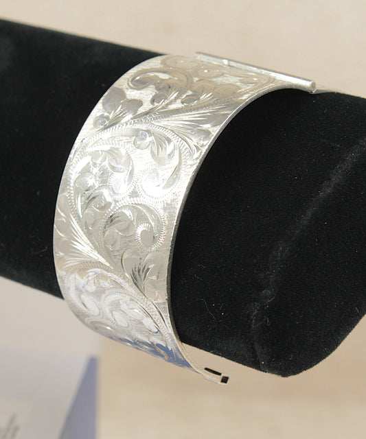 A wide silver cuff bracelet with intricate floral engraving, displayed on a black velvet jewelry stand against a light beige background.